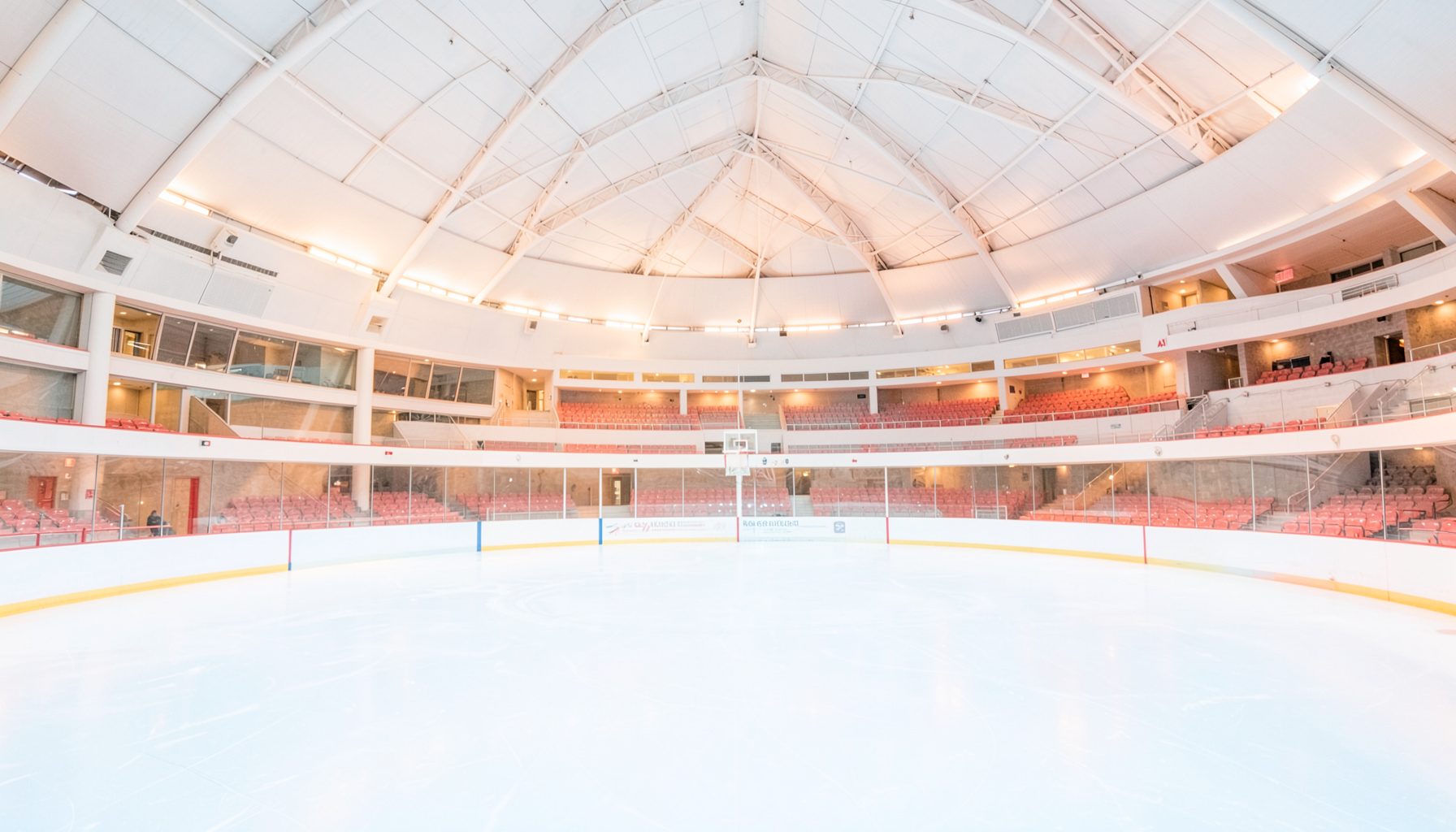 Wide view of a bright ice rink inside a modern arena with curved ceiling architecture, cool blue-white ice surface contrasting with warm amber arena lighting, empty and serene before a competition.
