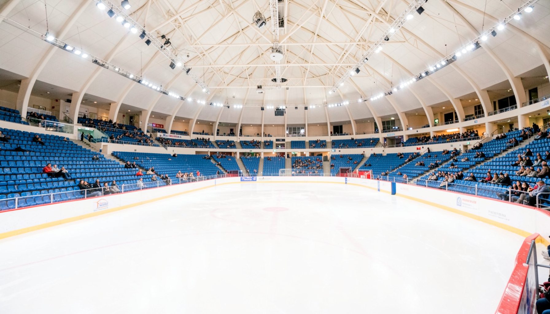 Wide view of the Palavela arena interior during a figure skating event, with rows of blue seating, bright overhead lights, and a pristine ice surface reflecting the warm glow of the arena. The atmosphere is calm and expectant before a session begins.