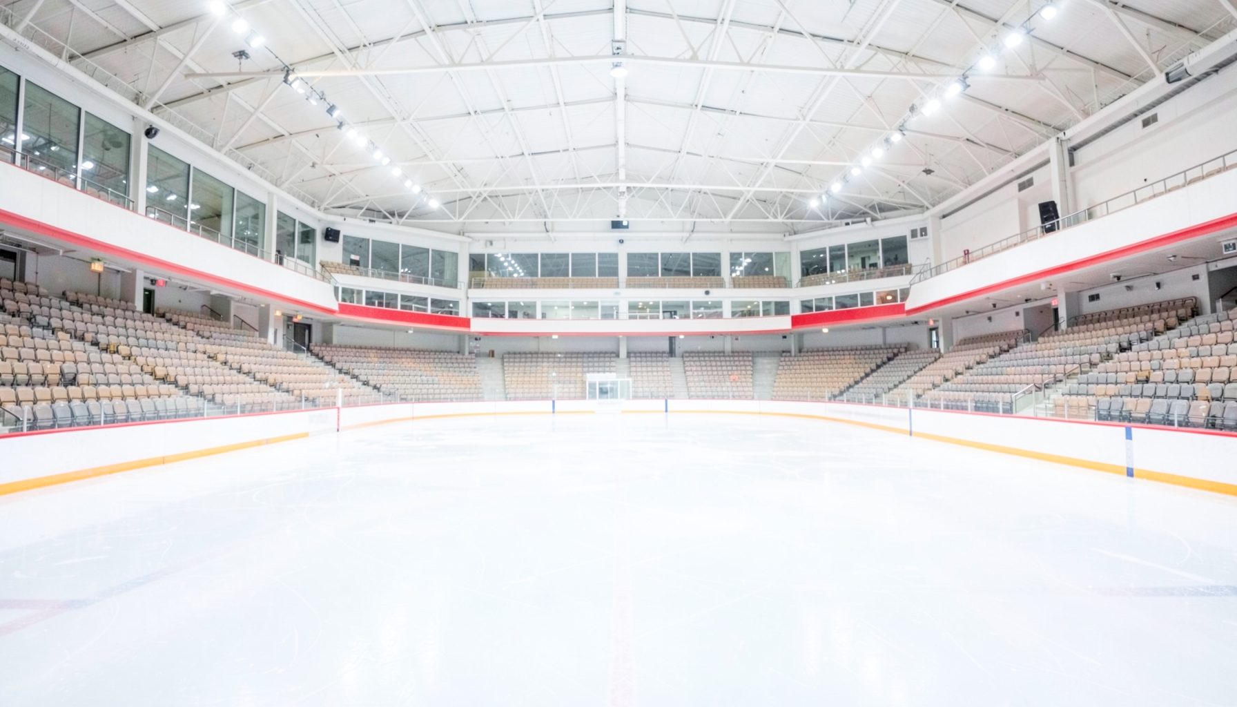 A wide, bright photograph of an empty ice rink inside a modern arena, with soft blue-white ice reflecting overhead lights and rows of empty seats in warm grey tones, conveying anticipation and elegance.