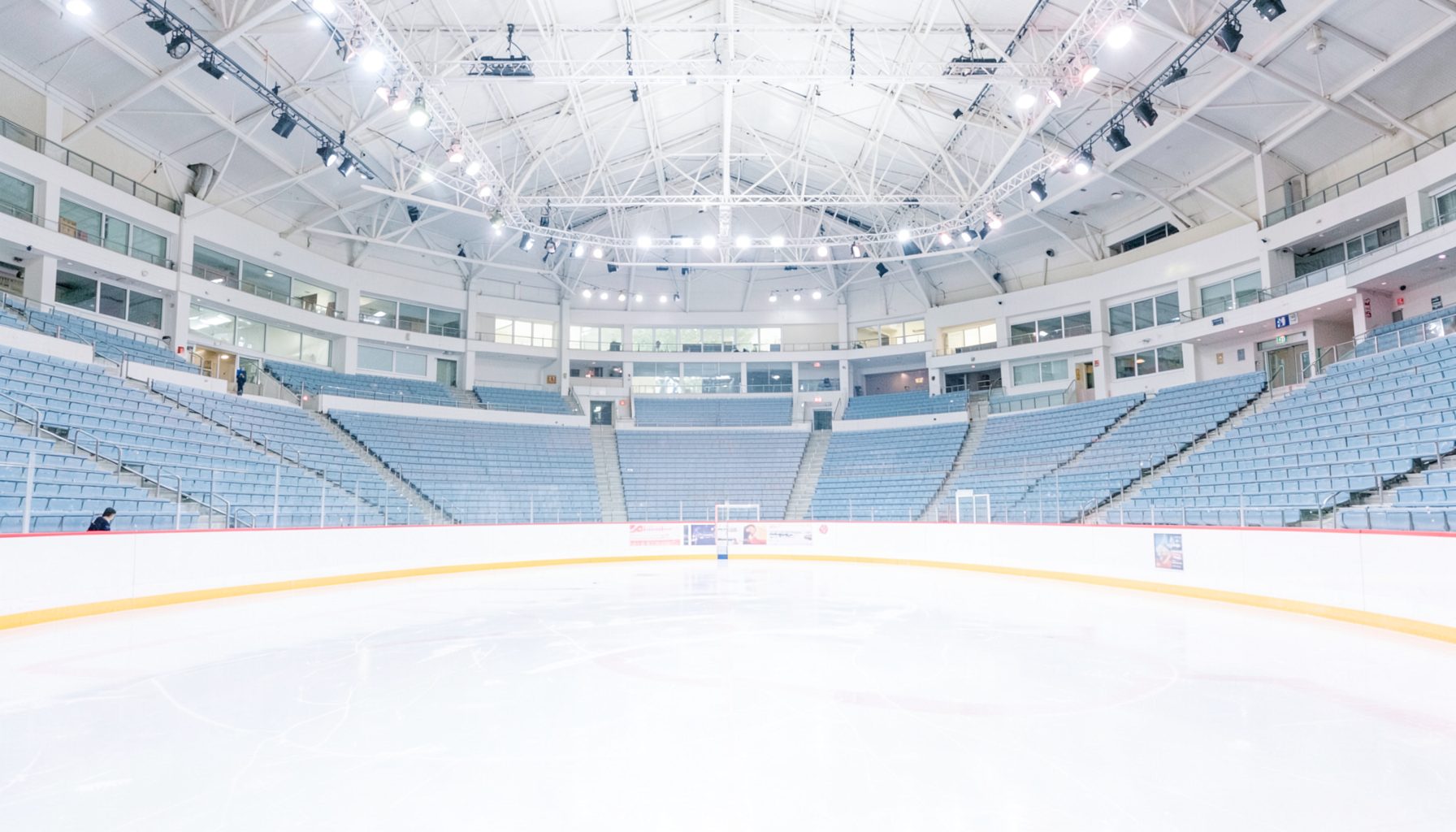 Wide view of a brightly lit ice rink inside a modern arena, with smooth ice reflecting overhead lights and empty tiered seating in soft blue-gray tones.